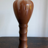 A brown plastic Japanese matcha whisk (chasuku) with a ribbed handle, standing on a dark wooden table against a plain off-white wall.
