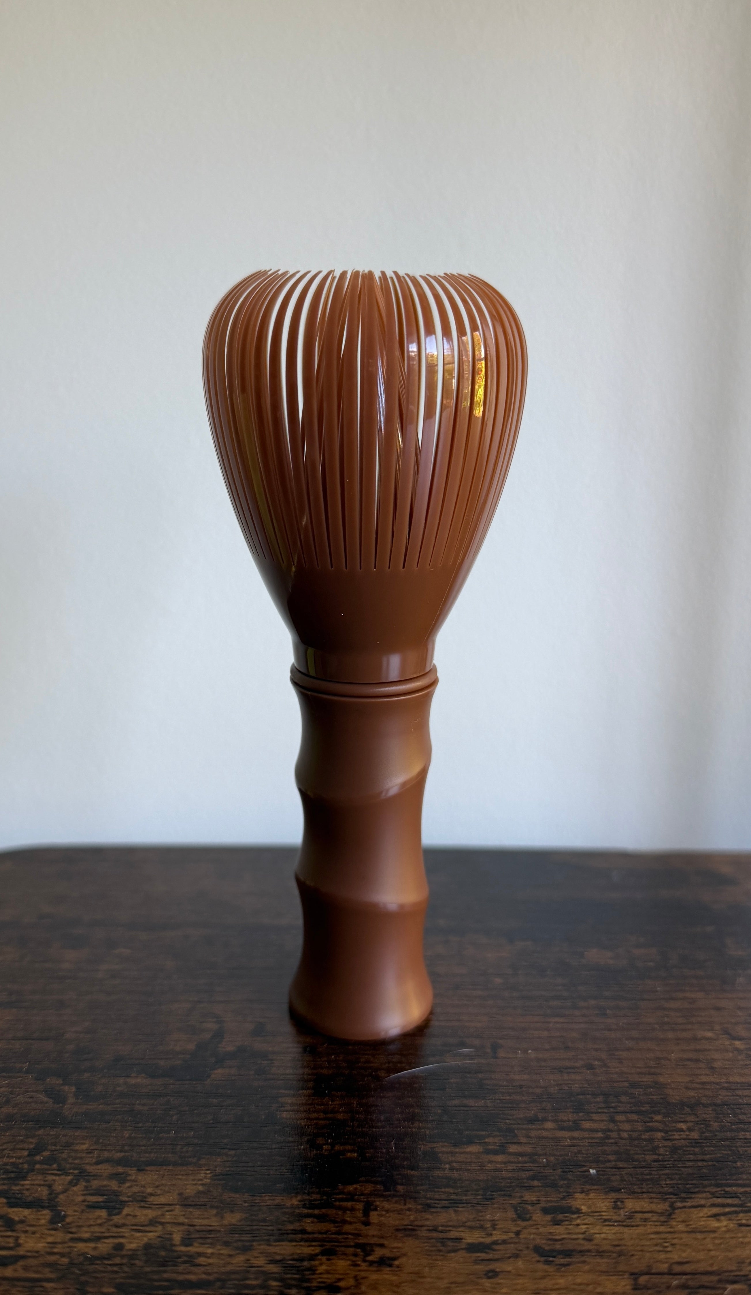 A brown plastic Japanese matcha whisk (chasuku) with a ribbed handle, standing on a dark wooden table against a plain off-white wall.
