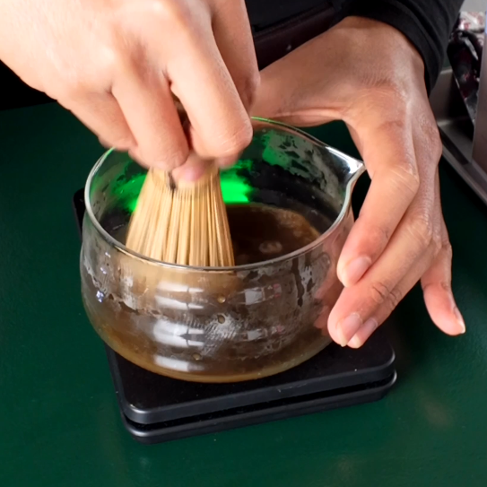 Person using a wooden whisk to stir a bowl of liquid on a green surface