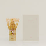 A matcha whisk with a gold-toned handle and bamboo bristles stands beside its cream-colored product box on a light beige background. The box has pink text reading 'Matcha Chasen Sandan Whisk' and 'Sakura Edition,' and the handle is printed with 'THEA.