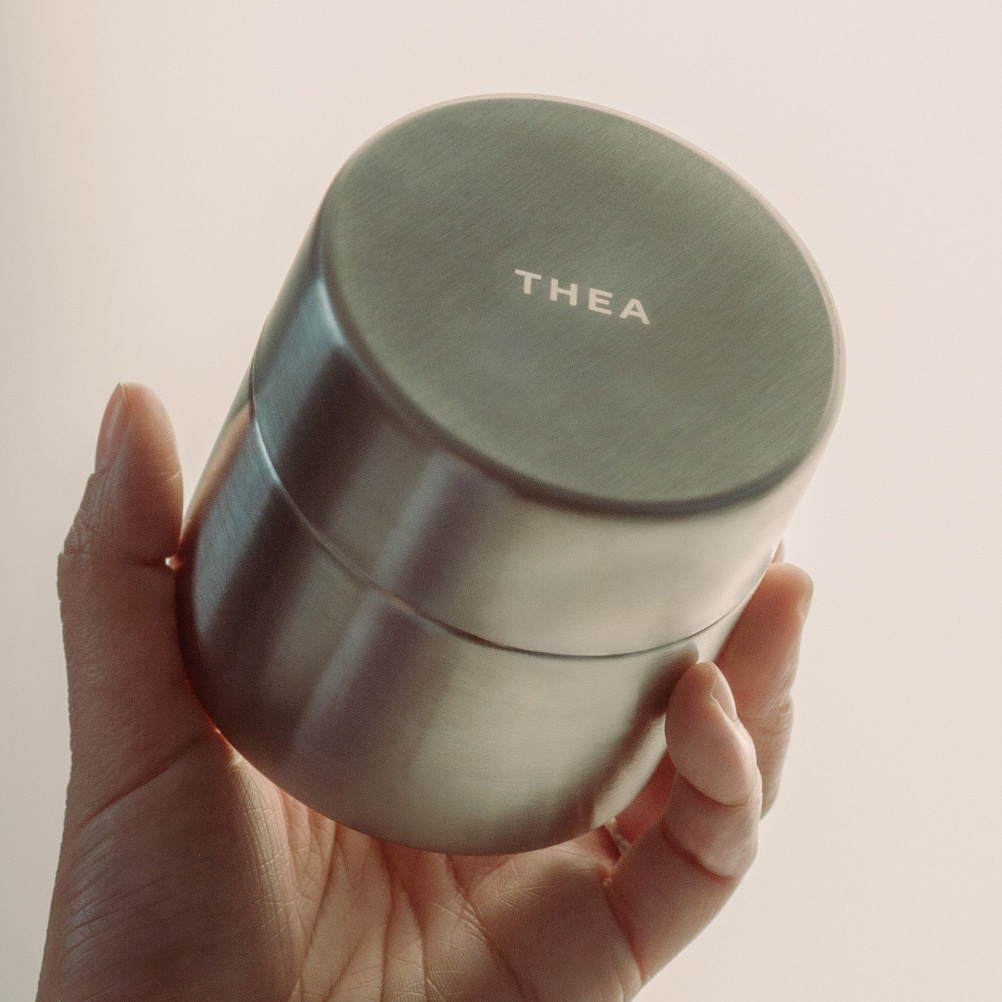A hand holding a brushed stainless steel cylindrical container with a lid, featuring the brand name 'THEA' printed on the lid, set against a solid light beige background.