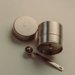 A stainless steel tea storage container with its lid off, shown next to a small metal measuring spoon on a neutral beige background. The brand name 'THEA' is embossed on the lid and engraved on the spoon handle.