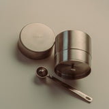 A stainless steel tea storage container with its lid off, shown next to a small metal measuring spoon on a neutral beige background. The brand name 'THEA' is embossed on the lid and engraved on the spoon handle.