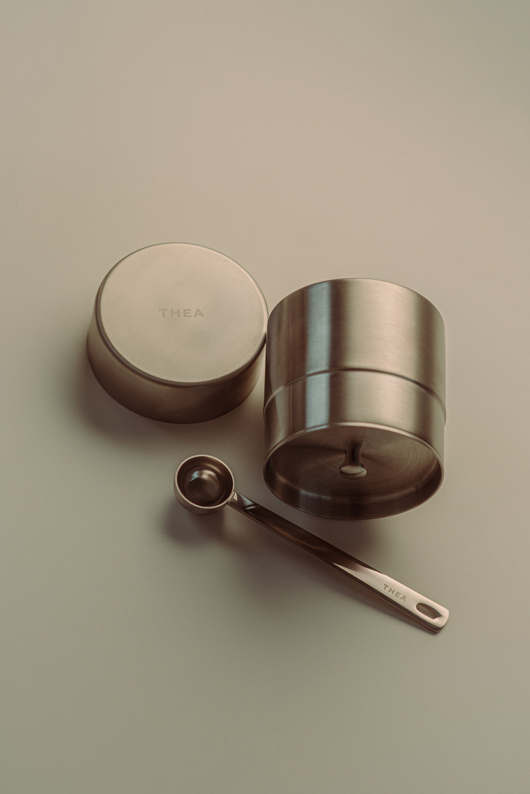 A stainless steel tea storage container with its lid off, shown next to a small metal measuring spoon on a neutral beige background. The brand name 'THEA' is embossed on the lid and engraved on the spoon handle.