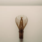 A traditional Japanese matcha whisk, also known as a chasen featuring a dark brown wooden handle and fine metallic gold wire prongs, standing upright on a neutral light gray surface.