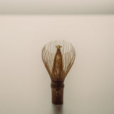 A traditional Japanese matcha whisk, also known as a chasen featuring a dark brown wooden handle and fine metallic gold wire prongs, standing upright on a neutral light gray surface.