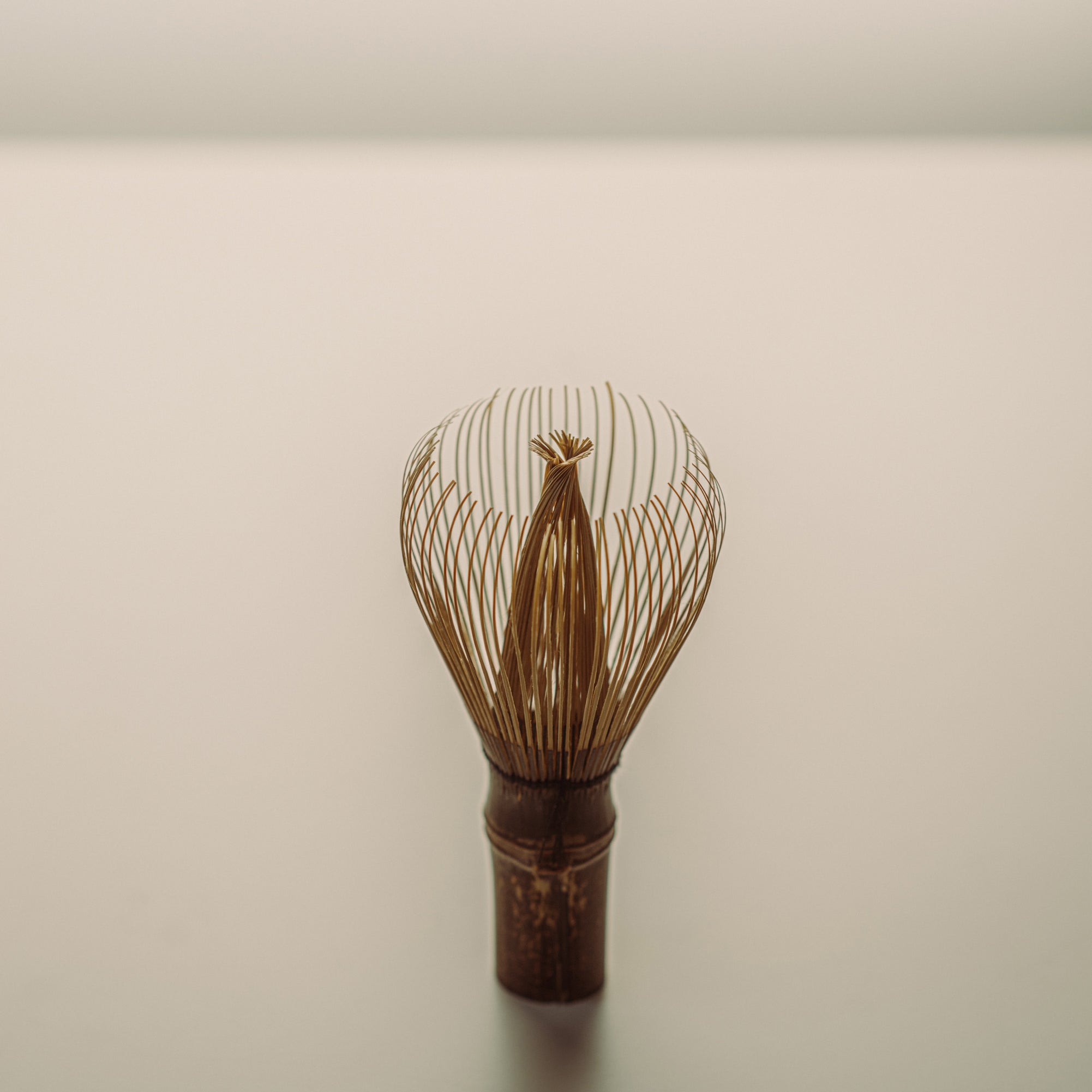 A traditional Japanese matcha whisk, also known as a chasen featuring a dark brown wooden handle and fine metallic gold wire prongs, standing upright on a neutral light gray surface.