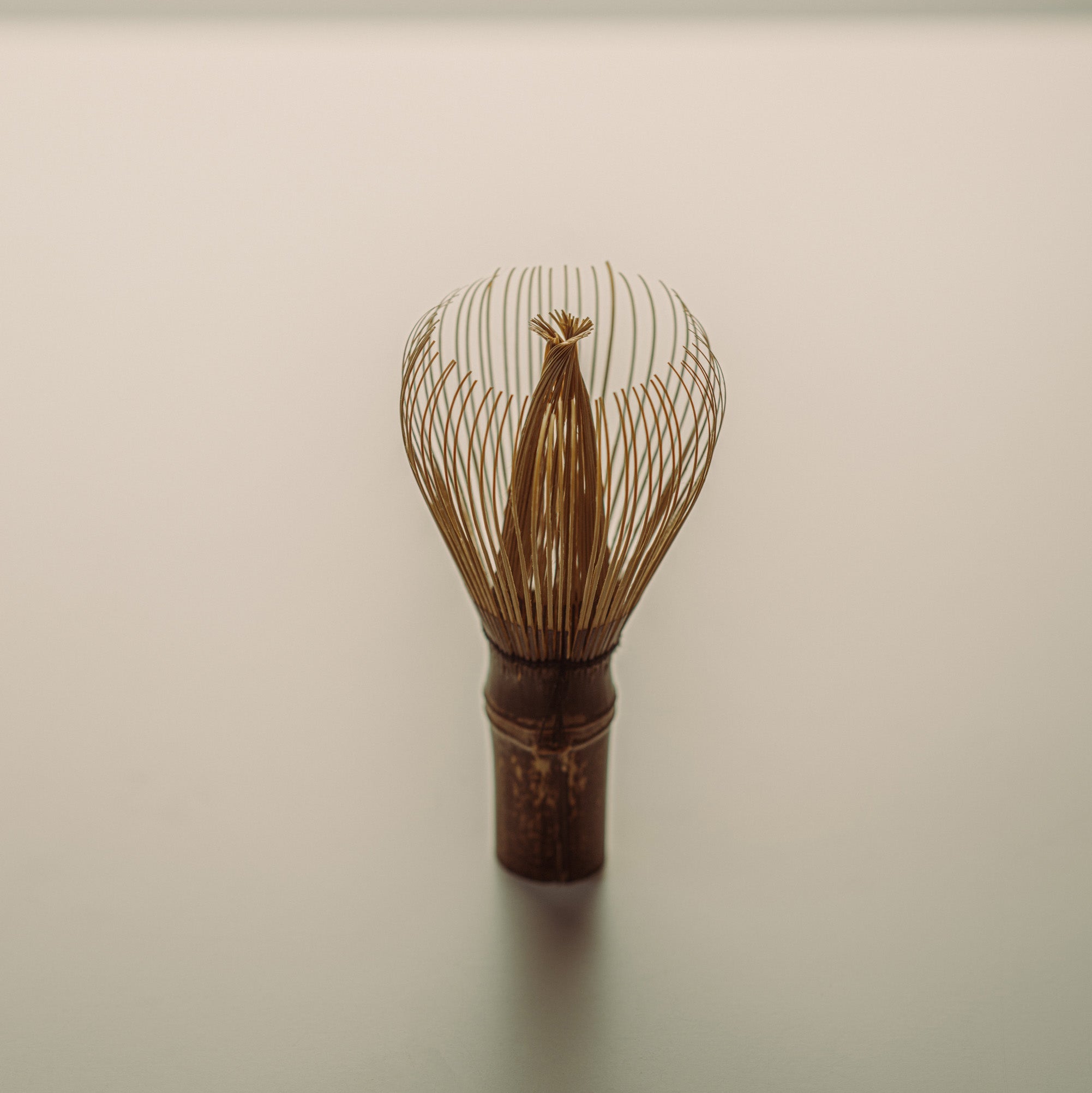 A traditional Japanese matcha whisk, also known as a chasen featuring a dark brown wooden handle and fine metallic gold wire prongs, standing upright on a neutral light gray surface.