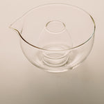 Clear glass mixing bowl with a pouring spout and a smaller inner bowl, shown on a plain light beige background.
