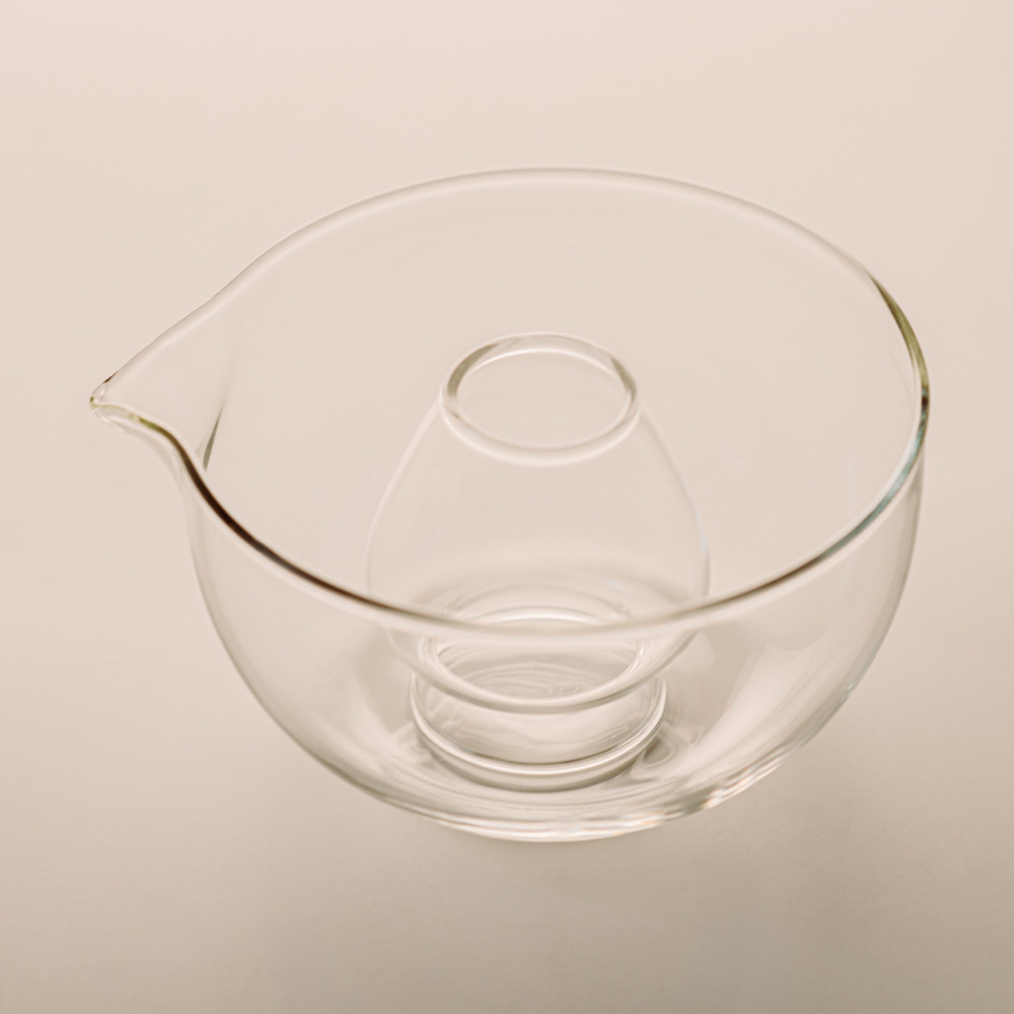 Clear glass mixing bowl with a pouring spout and a smaller inner bowl, shown on a plain light beige background.