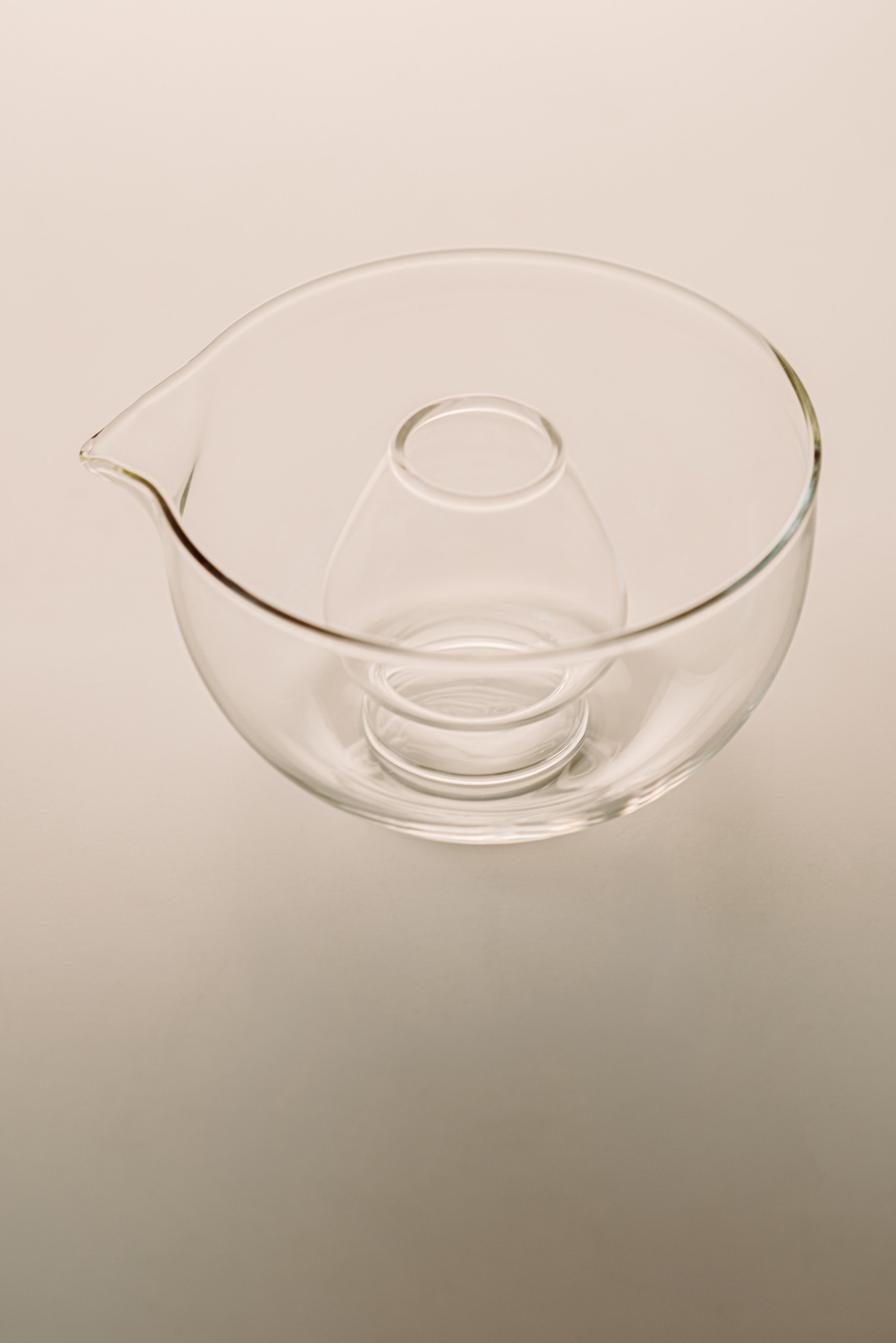 Clear glass mixing bowl with a pouring spout and a smaller inner bowl, shown on a plain light beige background.