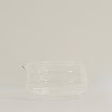 A clear glass pitcher with a small pouring spout, photographed against a plain light gray studio background.