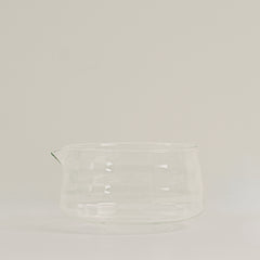 A clear glass pitcher with a small pouring spout, photographed against a plain light gray studio background.
