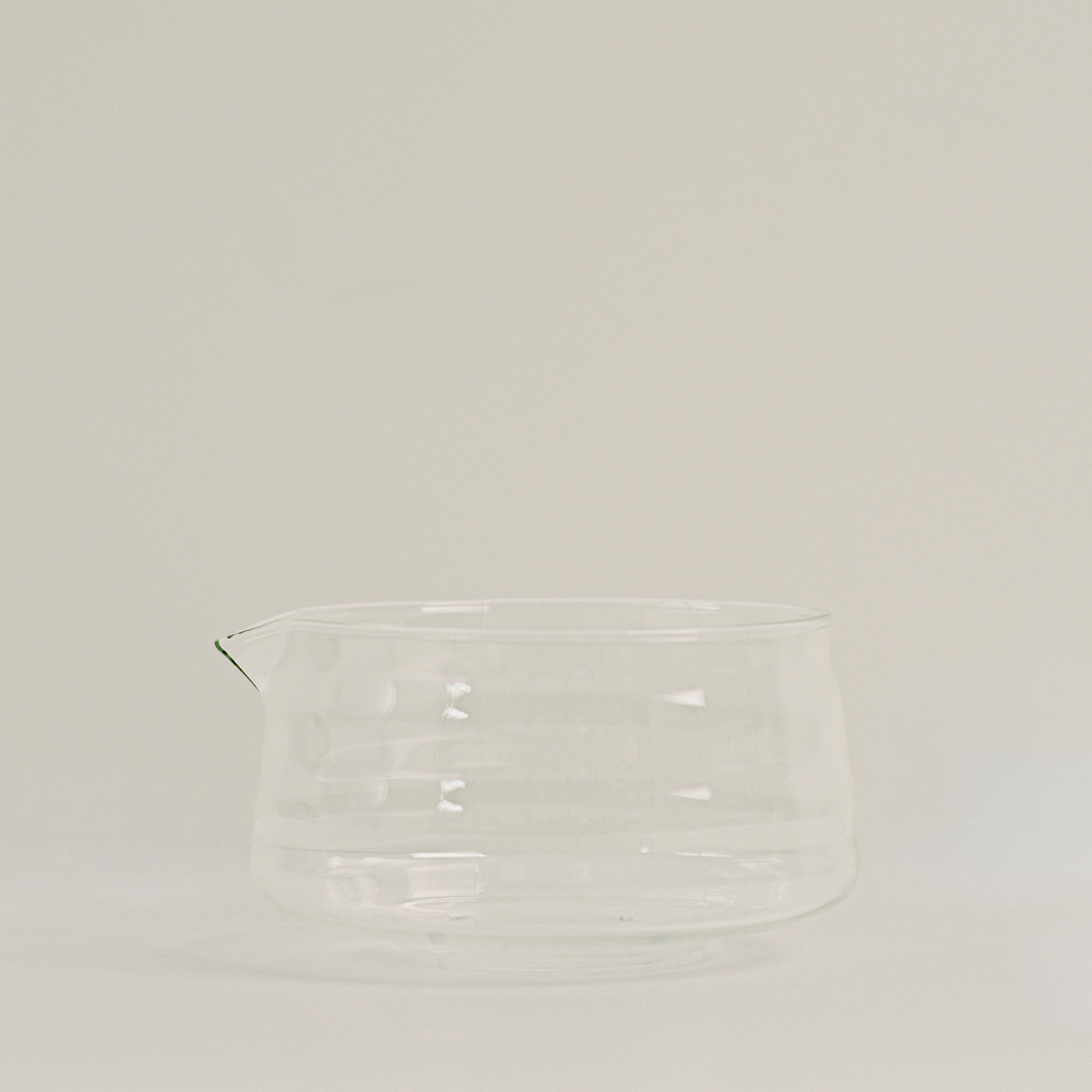 A clear glass pitcher with a small pouring spout, photographed against a plain light gray studio background.