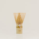 A traditional Japanese bamboo matcha whisk with a light beige bamboo handle, a white flower charm, and a fan of fine natural bamboo tines, standing upright on a light beige surface against a neutral cream background.