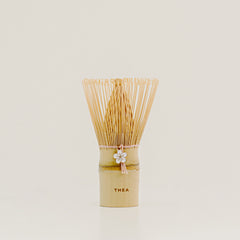 A traditional Japanese bamboo matcha whisk with a light beige bamboo handle, a white flower charm, and a fan of fine natural bamboo tines, standing upright on a light beige surface against a neutral cream background.