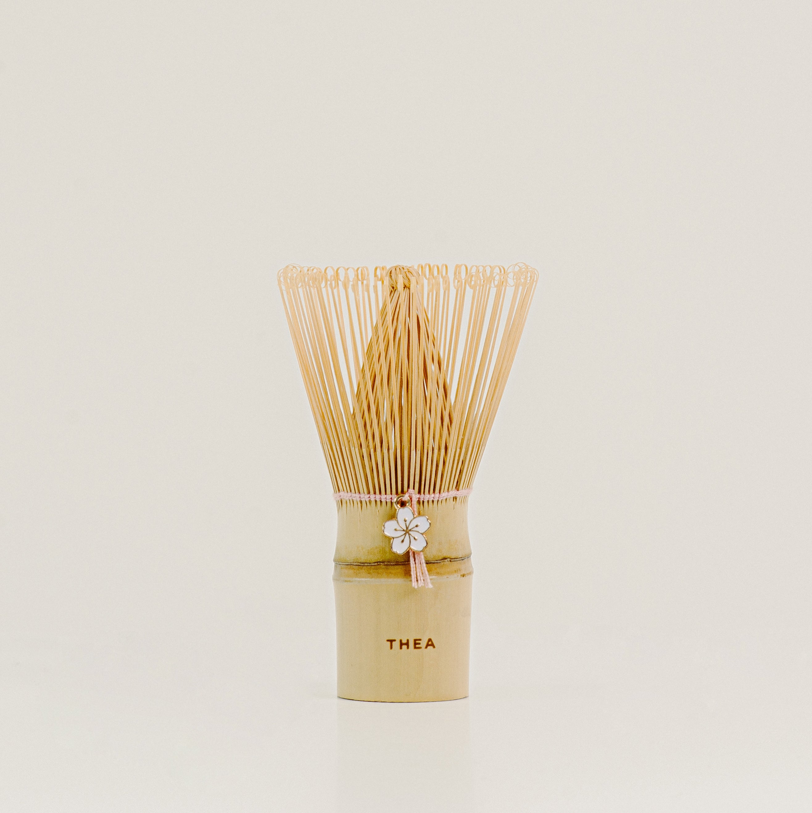 A traditional Japanese bamboo matcha whisk with a light beige bamboo handle, a white flower charm, and a fan of fine natural bamboo tines, standing upright on a light beige surface against a neutral cream background.