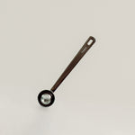 A single kitchen measuring spoon with a dark brown handle and a black bowl, featuring the brand name 'THEA' printed on the handle, set against a plain light gray background.