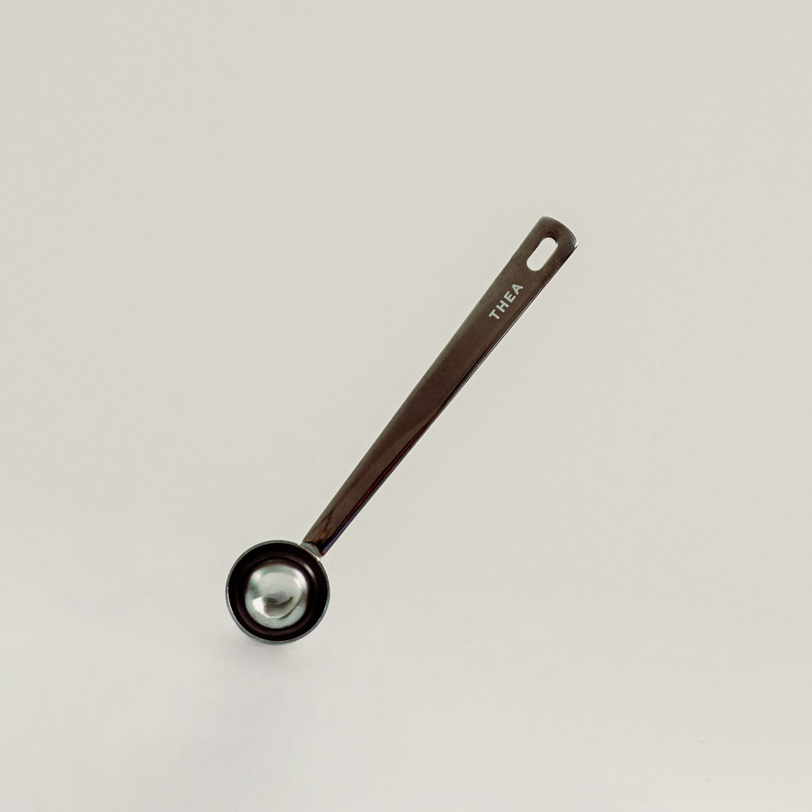 A single kitchen measuring spoon with a dark brown handle and a black bowl, featuring the brand name 'THEA' printed on the handle, set against a plain light gray background.