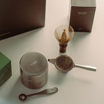 A tea preparation set arranged on a light surface, including a round metal container with the word 'THEA' on top, a whisk in a holder, a small strainer, a spoon, and dark brown boxes in the background.