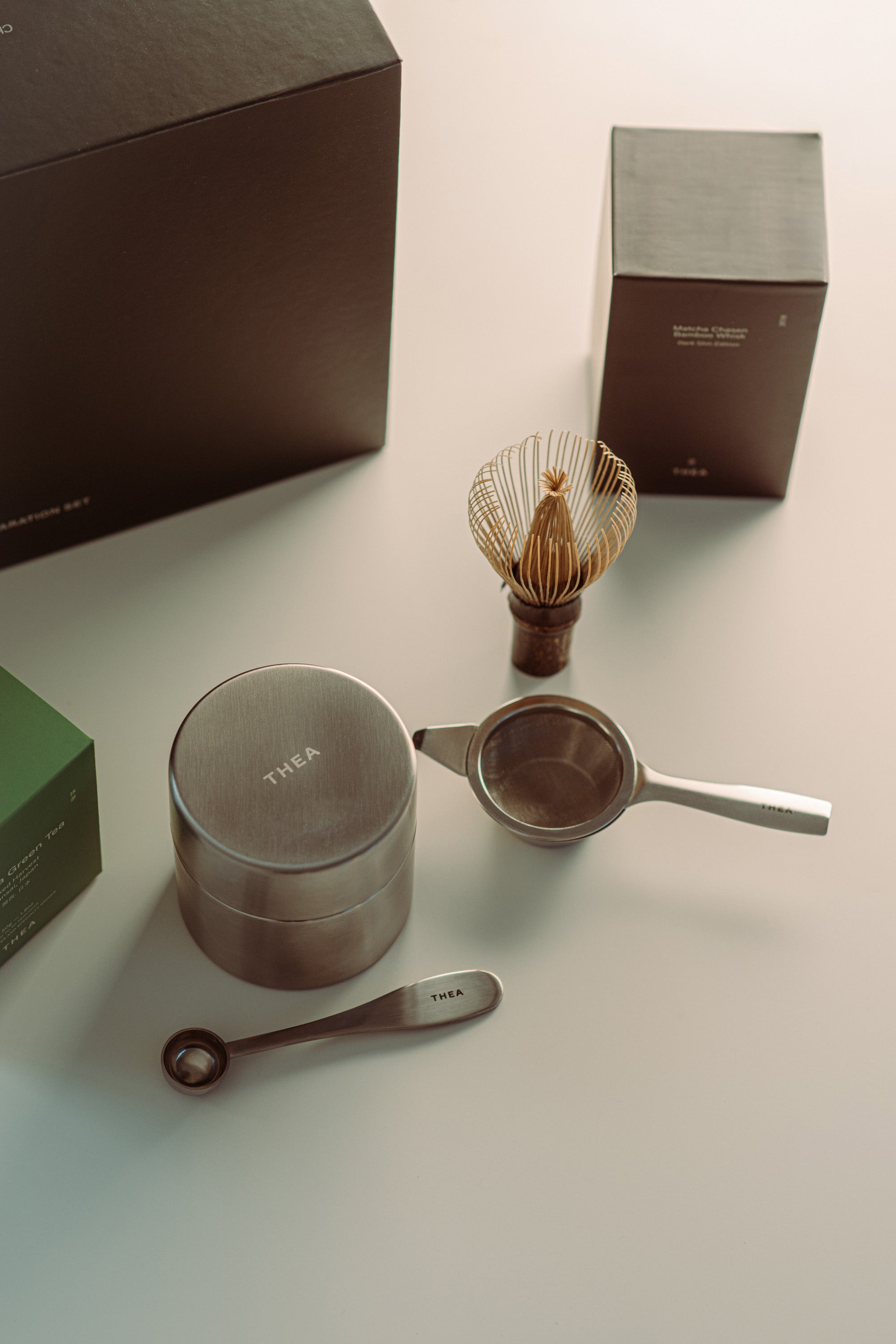A tea preparation set arranged on a light surface, including a round metal container with the word 'THEA' on top, a whisk in a holder, a small strainer, a spoon, and dark brown boxes in the background.