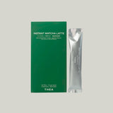 A green rectangular box labeled 'INSTANT MATCHA LATTE' stands next to a single silver sachet with matching text, on a plain light gray background. The box also shows 'THEA' near the bottom.